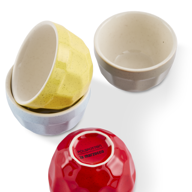 Four small cappuccino cups with faceted textured exteriors and smooth cream interiors, shown on a white background. One cappuccino cup is stacked inside another, one is upright showing its interior, one is upside down showing its red printed bottom, and one is standing upright showing its exterior.